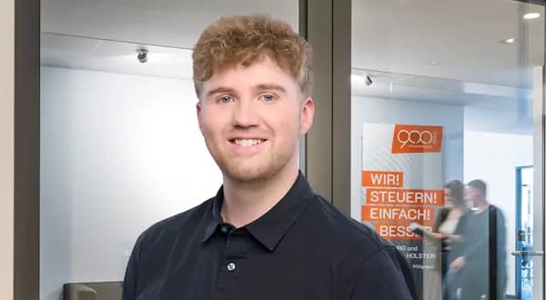Junger Mann mit lockigem blondem Haar lächelt vor einer Glaswand mit orange-weißem Firmenposter.