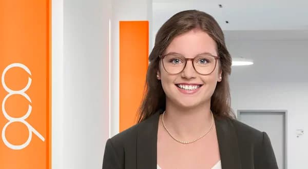 Junge Frau mit Brille und Perlenkette lächelt vor einem weißen Hintergrund mit orangefarbenen Streifen.